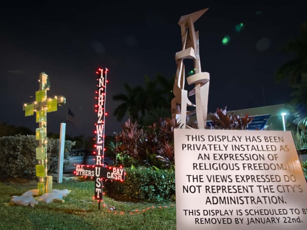 On the lawn of Hallandale Beach City Hall, a nighttime view of a private religious display featuring a cross with notes and lights, a sign reading "IN GOD WE TRUST ALL OTHERS PAY CASH," and a notice from the city explaining its purpose and scheduled removal date.