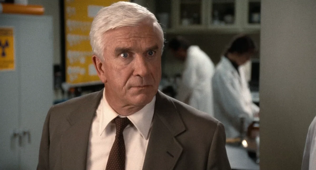 Older man in suit looks surprised in a lab setting, surrounded by scientists in white coats, intense expression.